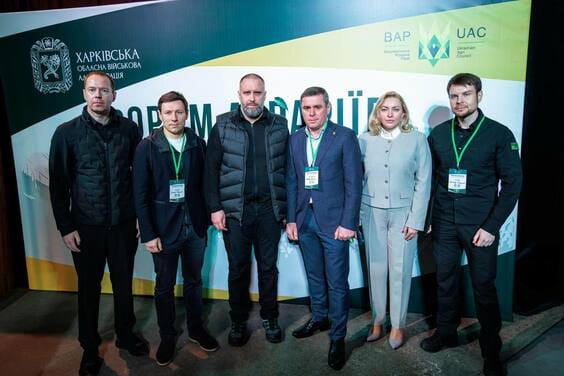 Шість осіб у діловитому вбранні стоять перед великим інформаційним стендом із офіційними логотипами та ідентифікаторами.