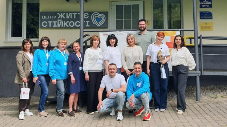 Група людей різного віку та в різноманітному одязі стоять та сидять біля будівлі із таблицею 