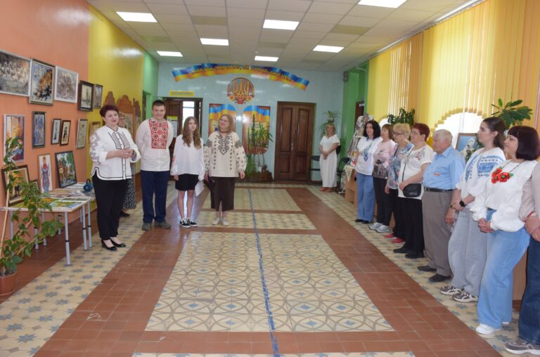 Група людей у вишиванках і національних вбраннях стоїть у коридорі з виставкою картин та кімнатними рослинами, утворюючи півколо та зібравшись біля однієї зі стін приміщення.