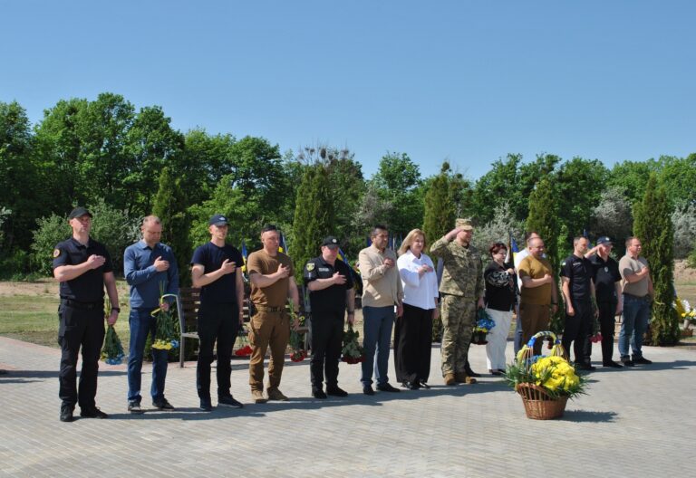 Група людей різного віку та в різному одязі стоїть на вулиці, приклавши праву руку до серця та салютуючи, біля кошика з квітами в очікуванні офіційної частини заходу.