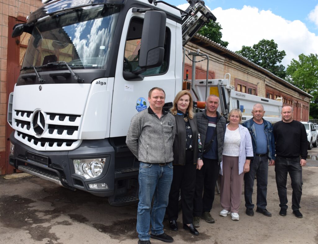Велика біла вантажівка Mercedes з логотипом alga біля будівлі, поруч з якою стоять шість людей.