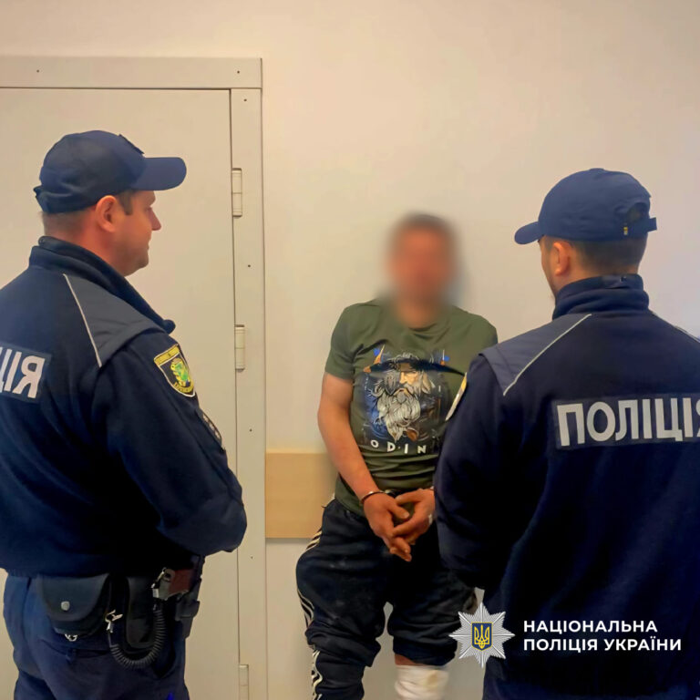 Два поліцейських у формі стоять по обидва боки від затриманого в зеленому одязі, який притулився до стіни із закованими у наручники руками.