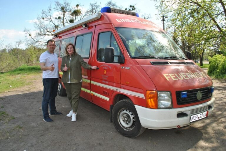 Чоловік і жінка показують великі пальці поруч із червоним автомобілем рятувальної служби з написом FEUERWEHR.