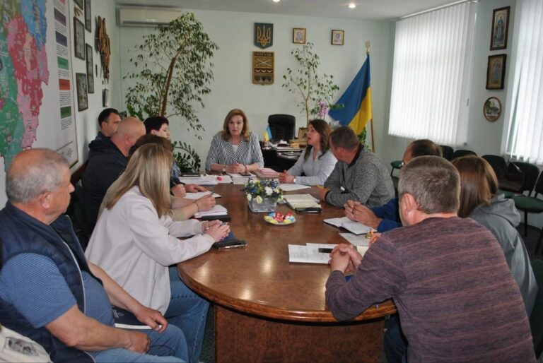 Кілька людей сидять навколо великого овального столу в кімнаті з українським прапором та різними прикрасами на стінах і займаються обговоренням документів.
