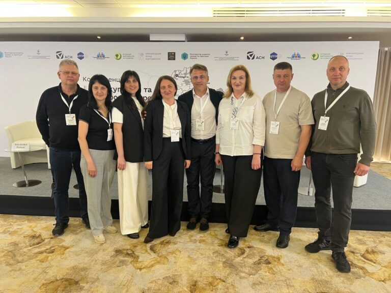 Група із семи осіб, переважно у діловому вбранні, посміхається перед камерою на тлі банера із зазначенням назви заходу українською мовою та різними логотипами організацій.