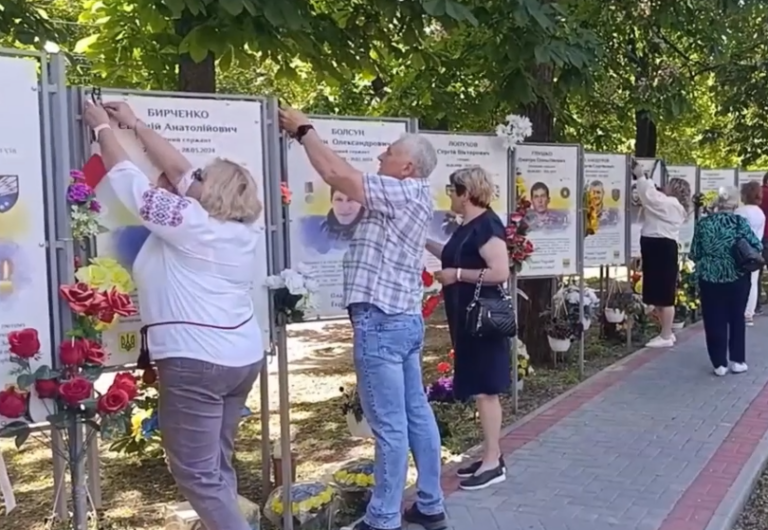 Жінка та чоловік прикріплюють квітку до меморіальної дошки із прізвищем і датами на ім'я Бірченко біля інших подібних стендів із квітами та світлинами вздовж доріжки.