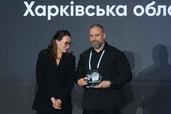 Чоловік та жінка, в чорних костюмах, стоять на тлі напису українською мовою; чоловік тримає в руках скляну статуетку.