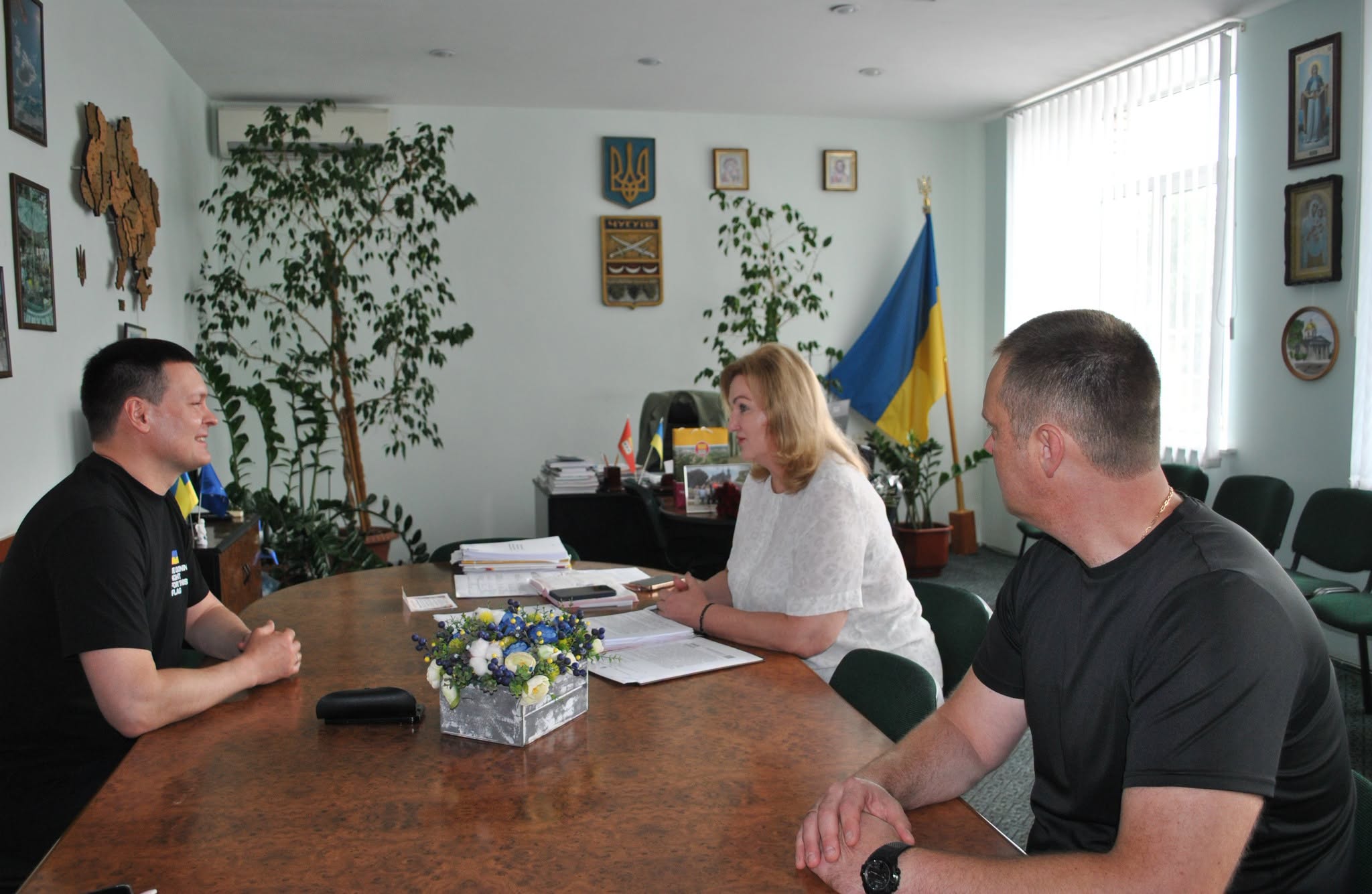 Жінка сидить між двома чоловіками за великим столом в офісній кімнаті, оточена українськими прапорами та різними прикрасами на стінах.