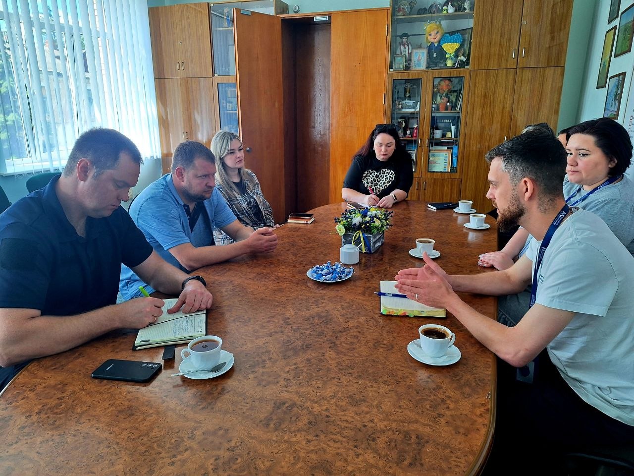 Кілька людей сидять навколо овального столу в кімнаті з дерев’яними шафами та вікном із вертикальними жалюзі; вони розмовляють та слухають виступ одного з учасників наради.