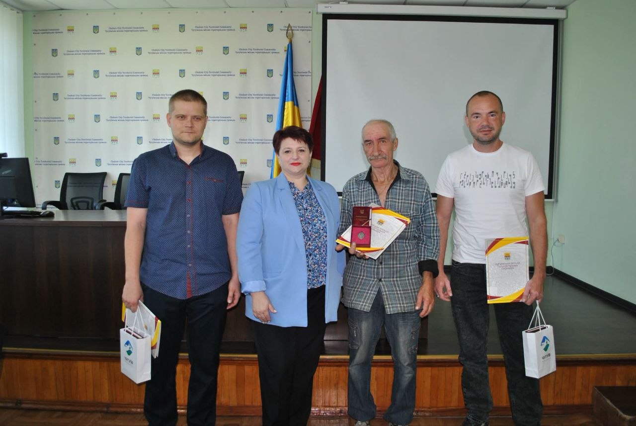 Четверо людей стоять поряд, посміхаючись; серед них жінка у світло-блакитному піджаку та троє чоловіків, двоє з яких тримають пакети, а один — нагороду у вигляді медалі на подіумі та сертифікати.