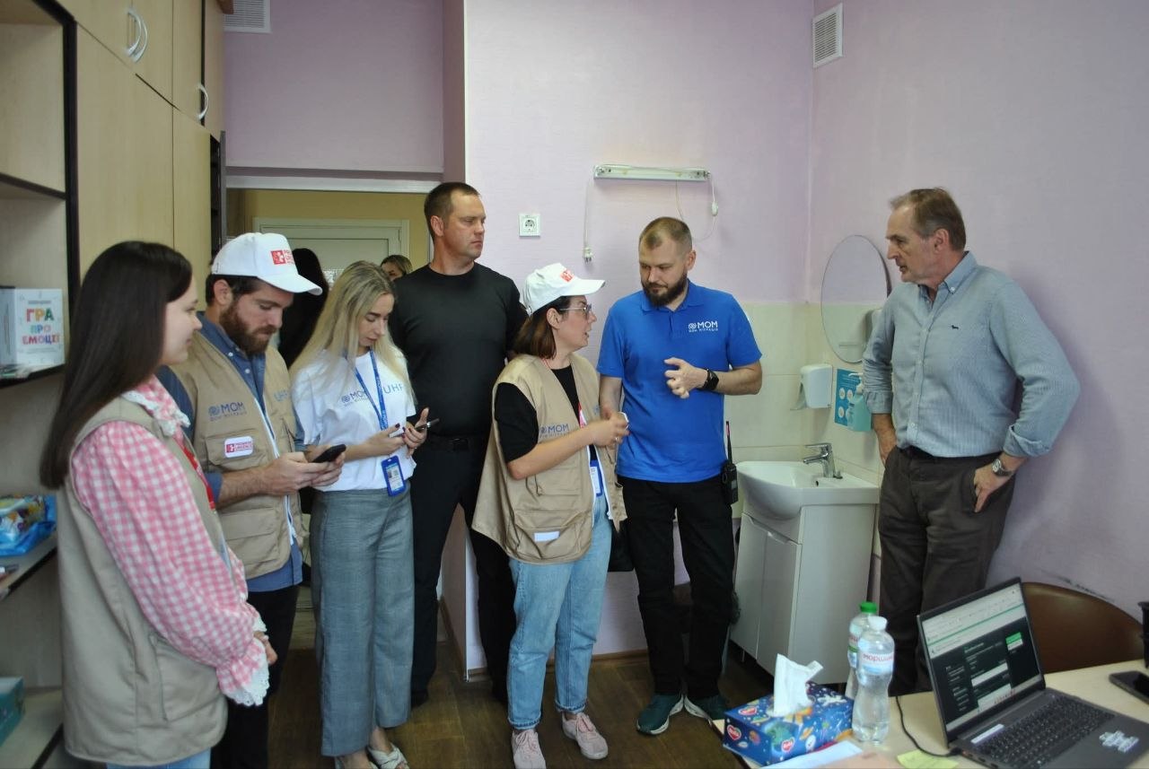Кілька людей в уніформі та цивільному одязі зібрались у кімнаті біля раковини, розглядаючи інформаційний пристрій із відкритим інтернет-ресурсом.