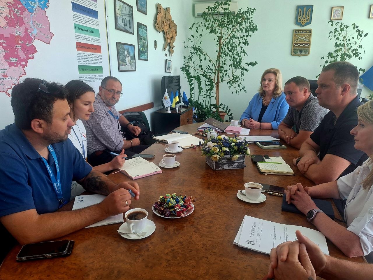 Кілька осіб сидять за столом у кімнаті, навколо якого розташовані папери та чашки кави; вони, імовірно, беруть участь у нараді.