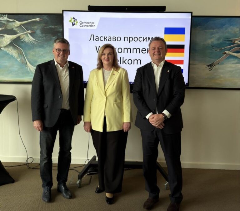 Три людини в офіційному одязі стоять перед екраном із привітальною інформацією українською мовою та прапорами Нідерландів і Німеччини.