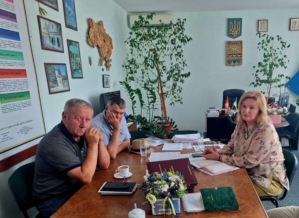 Троє людей сидять за столом в офісі, серед документів та квітів, жінка праворуч дивиться на глядача, а двоє чоловіків ліворуч виглядають задумливими.