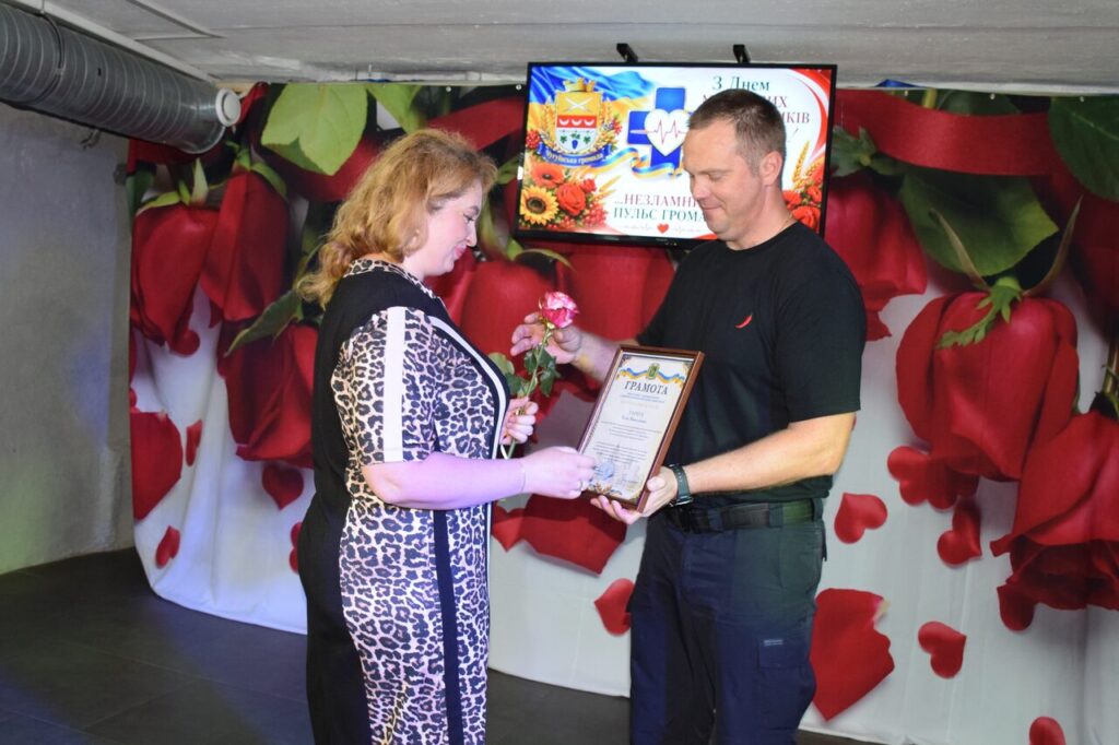 Чоловік вручає жінці в леопардовій сукні рамку з грамотою та квітку на тлі екрана з написом 
