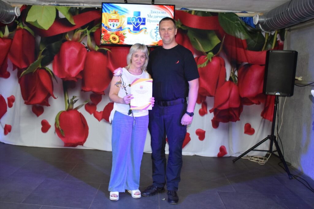 Жінка з грамотою та чоловік стоять на сцені перед фоном з червоними трояндами та екраном із написом 