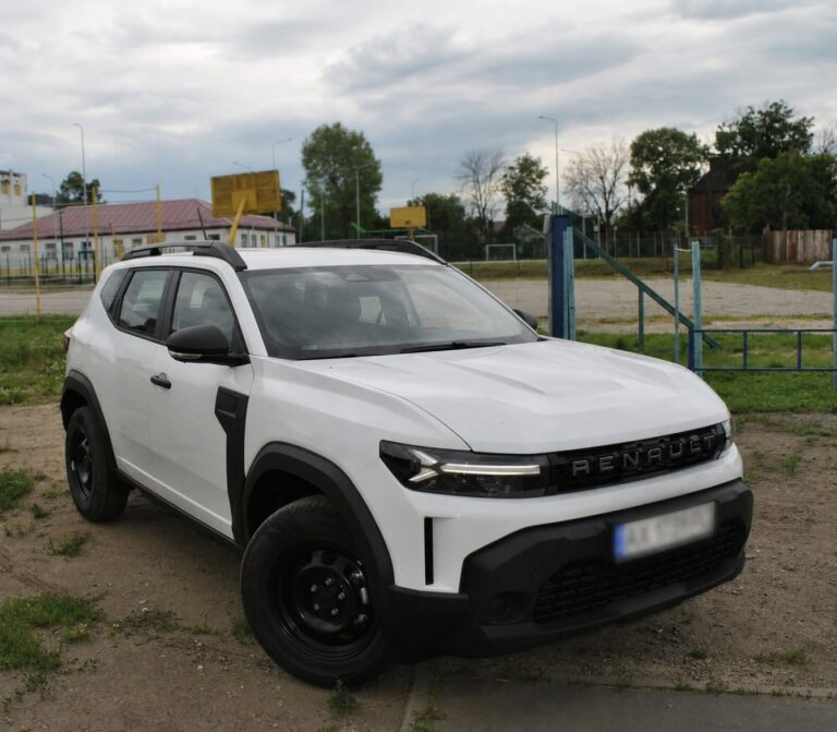 Білий автомобіль Renault припаркований на ґрунтовій ділянці біля дитячого майданчика з обладнанням та металевою гіркою на задньому плані.