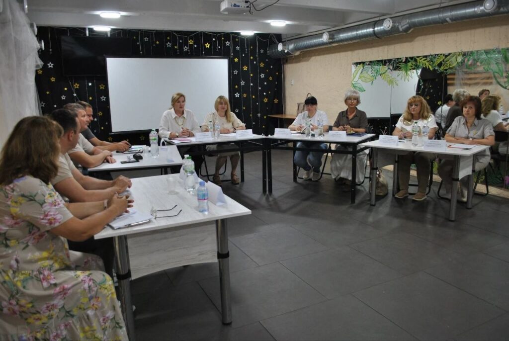 Кілька людей сидять за столами у приміщенні з білою дошкою та проектором на стелі.