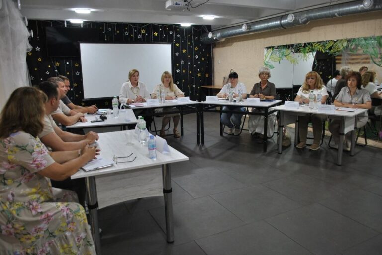 Кілька людей сидять за столами у приміщенні з білою дошкою та проектором на стелі.