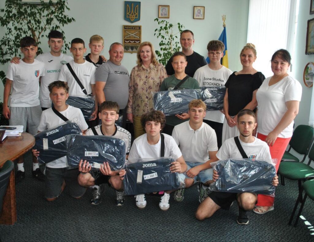 Група людей, переважно чоловіків, зображена в приміщенні, більшість із них тримають у руках упаковані чорні спортивні сумки Joma.