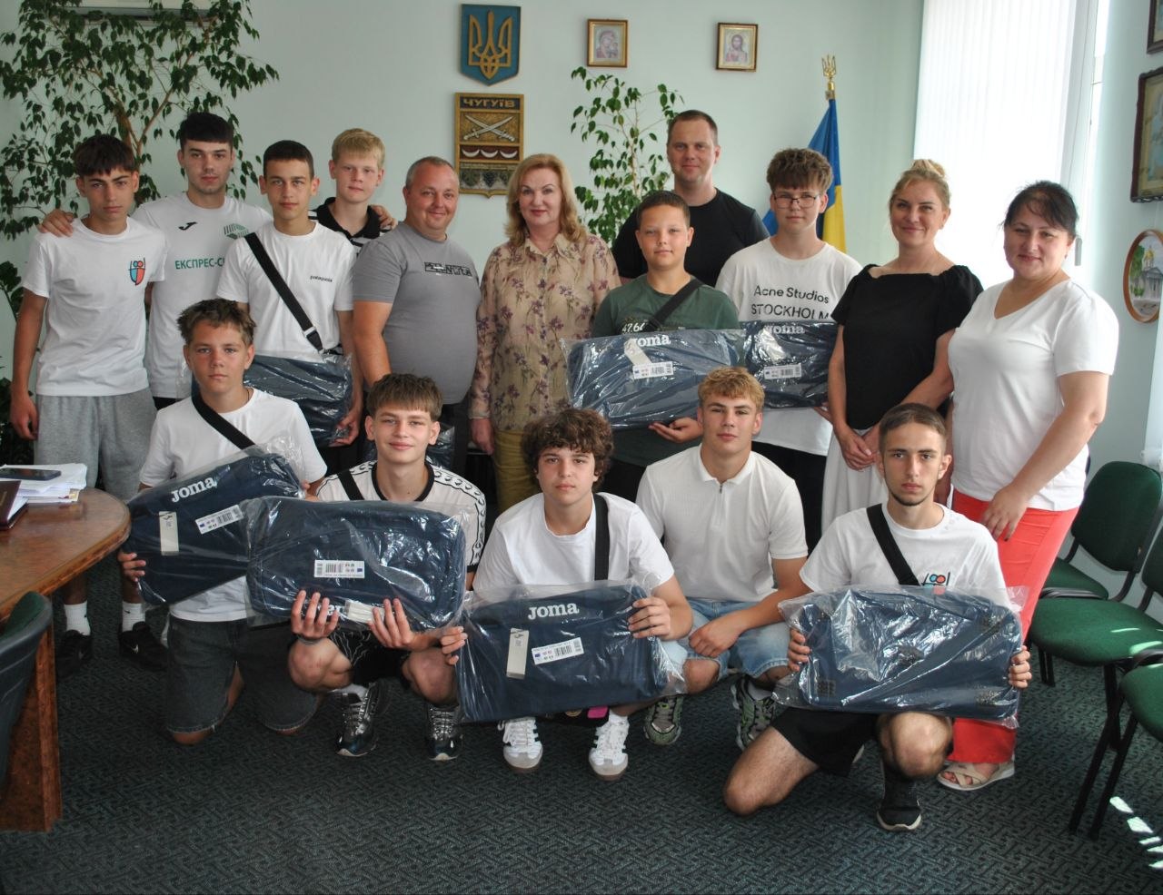Група людей, переважно чоловіків, зображена в приміщенні, більшість із них тримають у руках упаковані чорні спортивні сумки Joma.