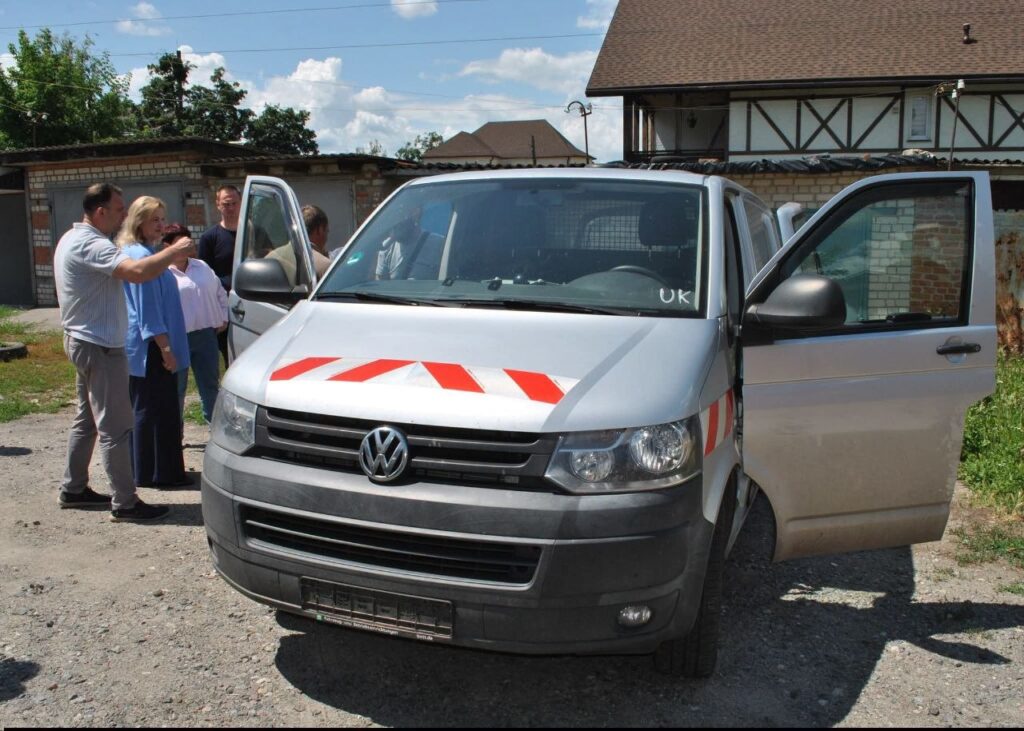 Сріблястий мікроавтобус Volkswagen з червоними смугами на капоті та відкритими дверима, біля якого стоять люди.