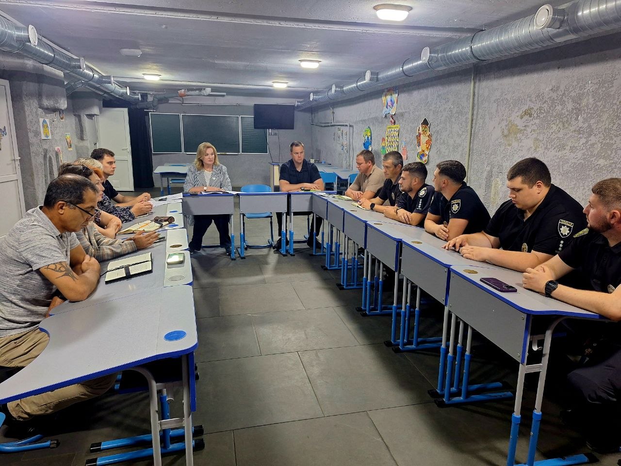 Жінка сидить за столом серед групи людей, які сидять півколом і уважно слухають її.