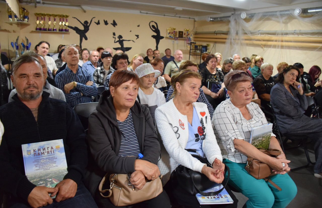 Група людей сидить у залі, а чоловік на передньому плані тримає книгу з назвою 