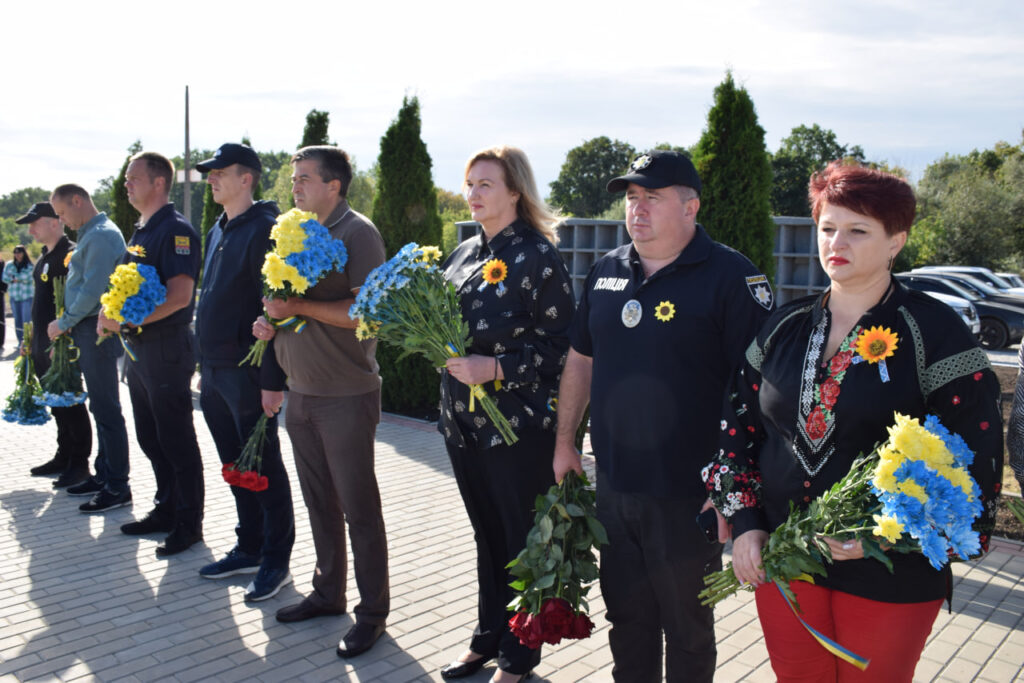 Група людей, серед яких поліцейські, стоїть у шеренгу, тримаючи букети синьо-жовтих та червоних квітів.