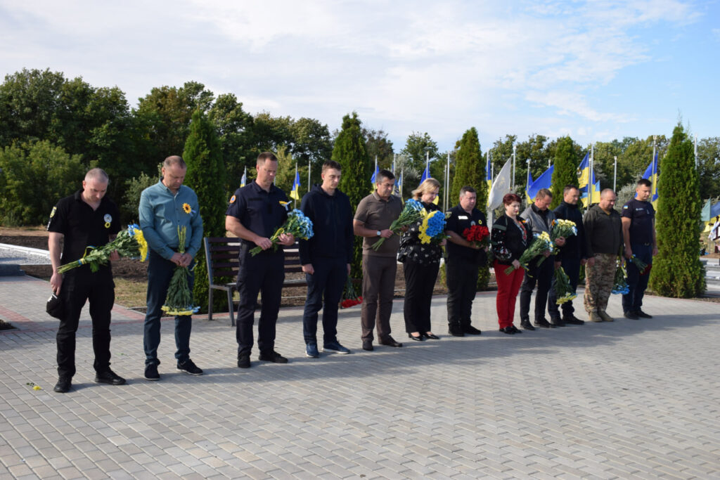 Група людей стоїть у шерензі, тримаючи букети квітів на тлі українських прапорів.