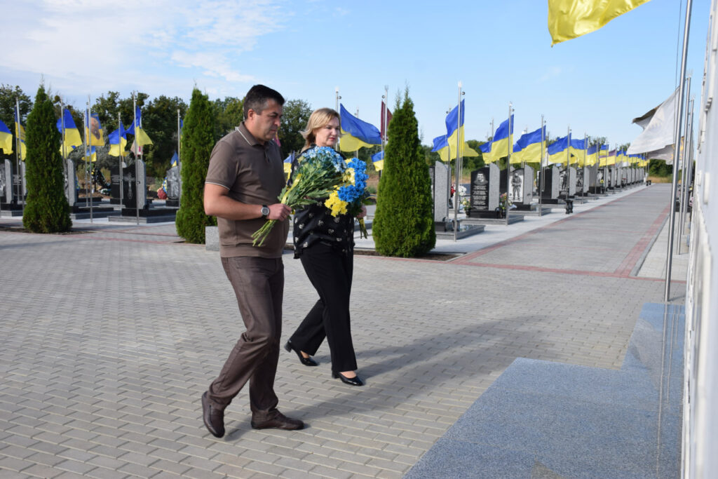 Чоловік і жінка йдуть алеєю, тримаючи букети синьо-жовтих квітів, на тлі ряду могил, прикрашених українськими прапорами.
