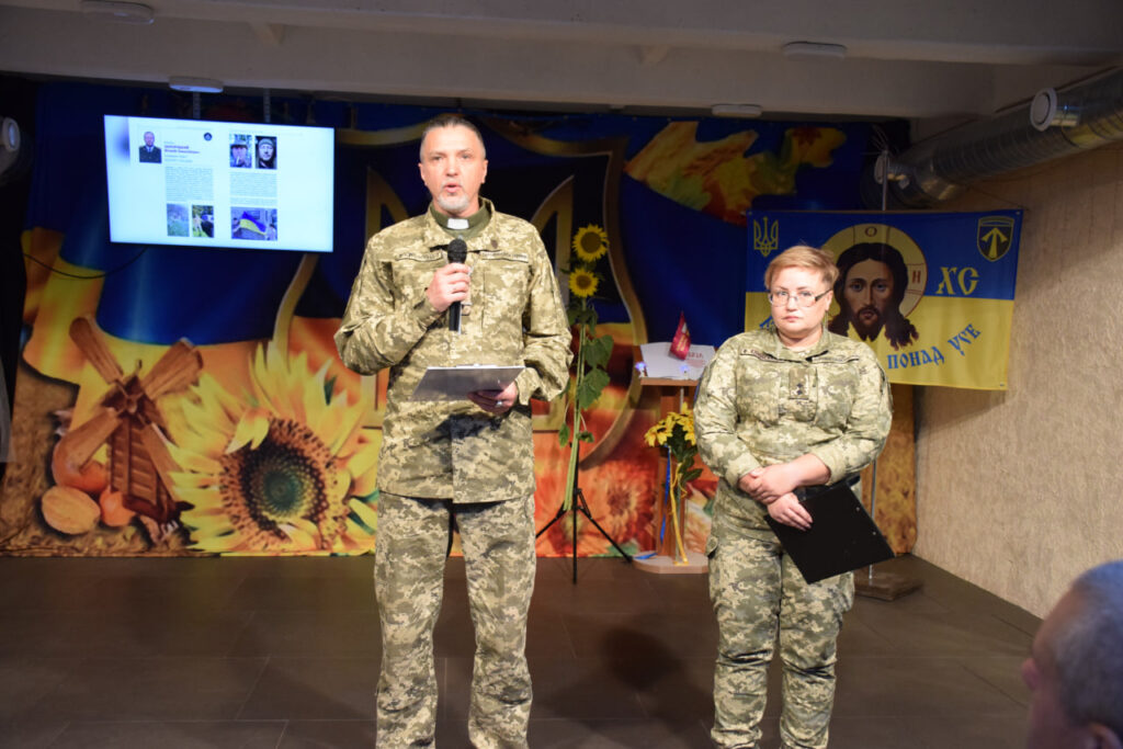 Чоловік у військовій формі з кліричним коміром говорить у мікрофон на сцені поруч із жінкою у формі, на тлі екрана з презентацією та релігійного прапора з образом Ісуса Христа.