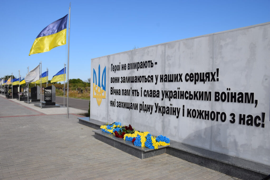 Меморіальна стіна з написом «Герої не вмирають - вони залишаються у наших серцях! Вічна пам'ять і слава українським воїнам, які захищали рідну Україну і кожного з нас!» та покладеними квітами.