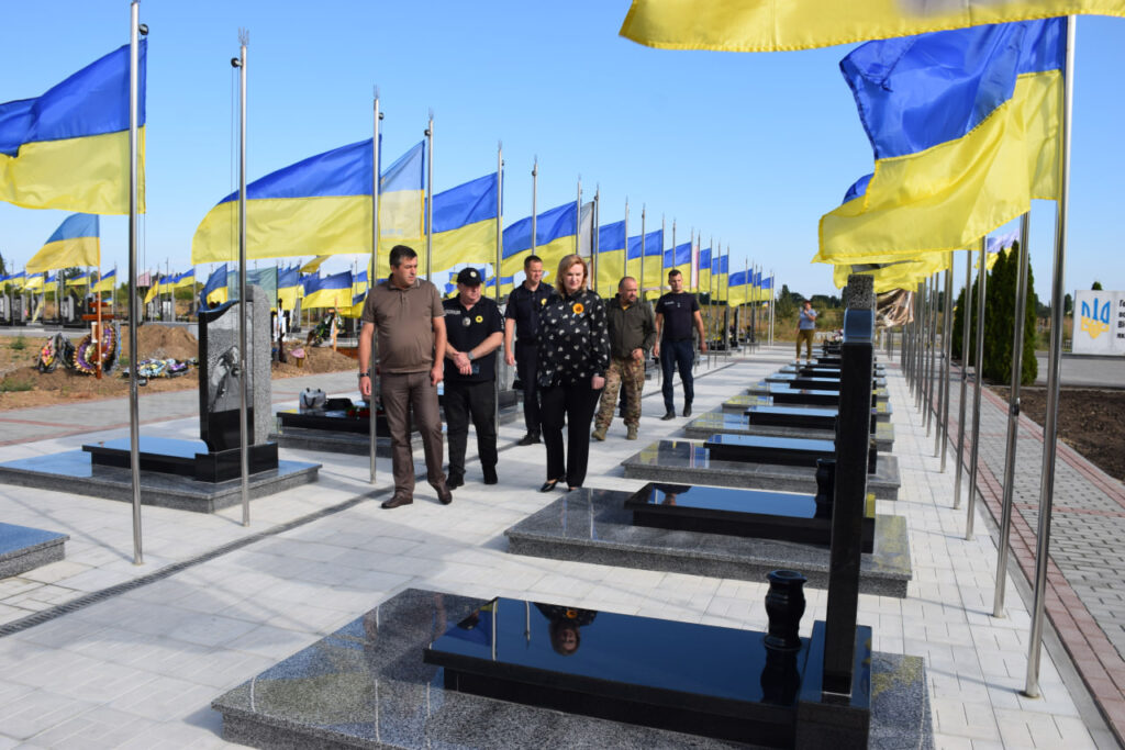 Люди проходять повз ряди чорних надгробків, біля кожного з яких встановлено прапор України.