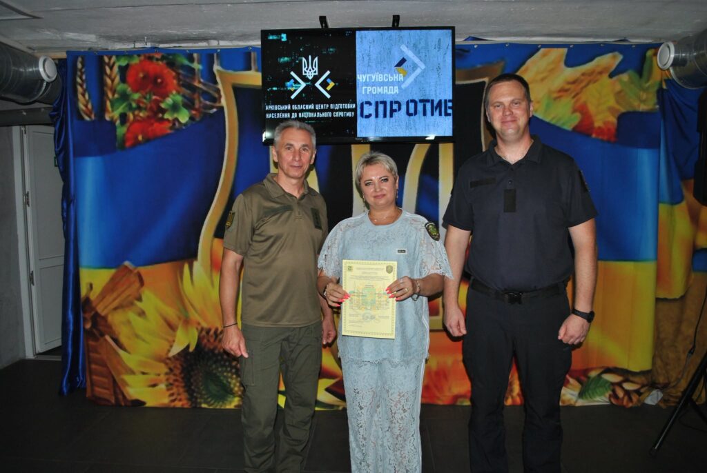Троє людей стоять перед банером із соняшниками та прапором України, де жінка в центрі тримає сертифікат, а на екрані вище відображено написи