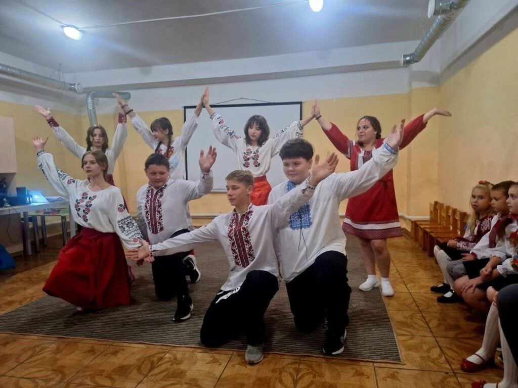Група дітей у традиційних вишиванках виконує народний танець у приміщенні.