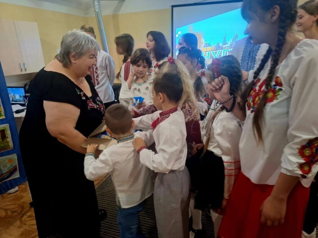 Final Polish:* Жінка в чорній сукні спілкується з групою дітей у вишиванках під час заходу.