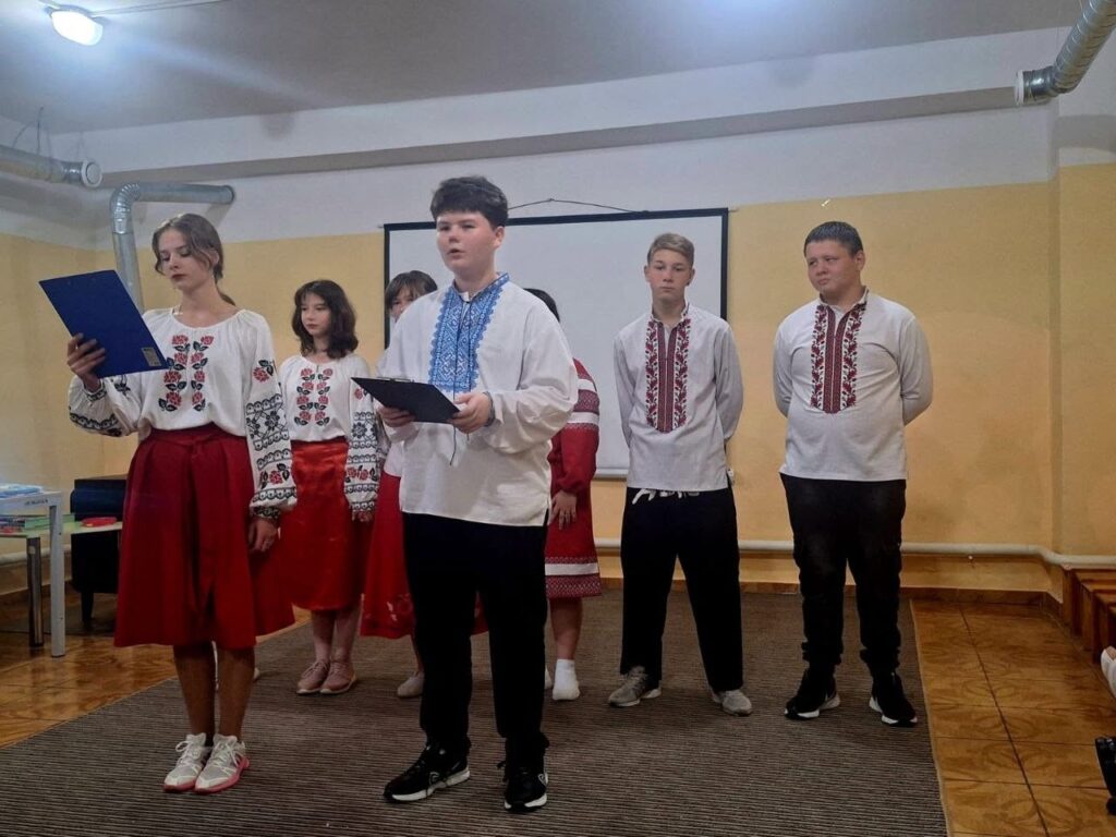 Група підлітків у традиційних вишиванках стоїть у залі та читає текст з папок.