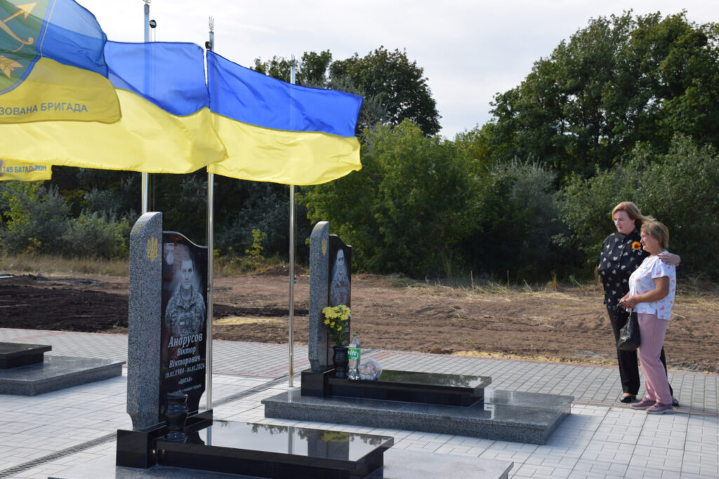 Чорний надгробок з портретом загиблого воїна Віктора Андрусова та датами його життя, поруч з яким хвилюються українські прапори, а дві жінки