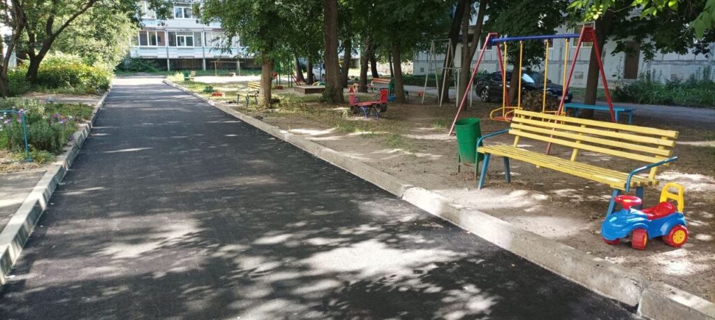 Асфальтована стежка в парку, поруч з якою розташовані жовта лавка, гойдалки та дитяча машинка.
