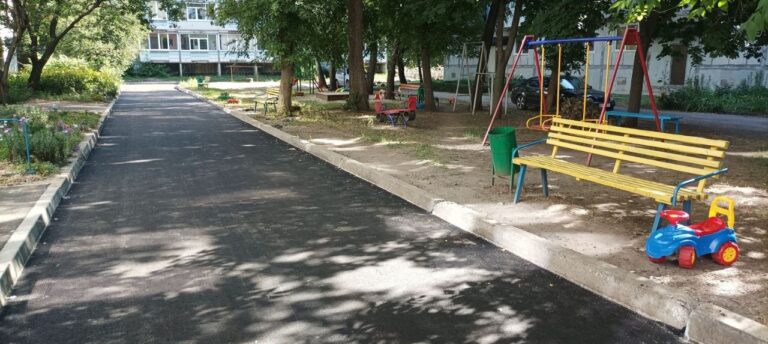 Асфальтована стежка в парку, поруч з якою розташовані жовта лавка, гойдалки та дитяча машинка.
