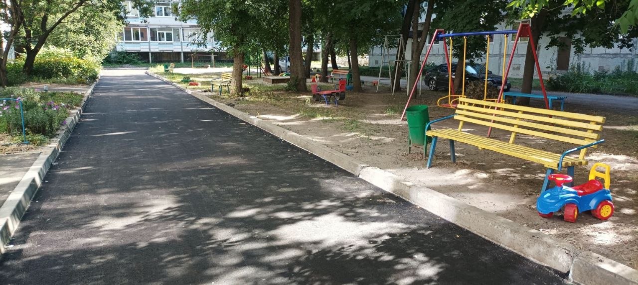 Асфальтована стежка в парку, поруч з якою розташовані жовта лавка, гойдалки та дитяча машинка.