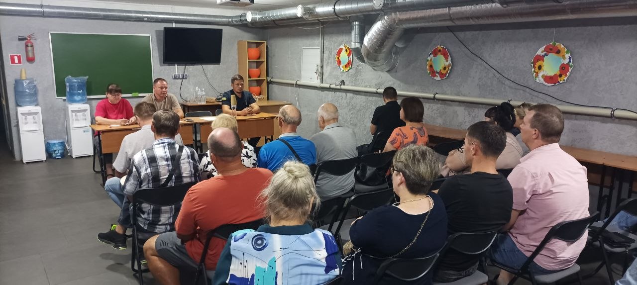 Група людей сидить на стільцях і слухає трьох спікерів, які сидять за столом у приміщенні з дошкою.