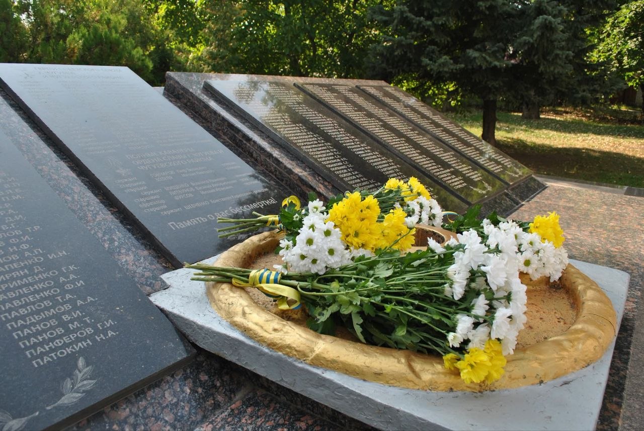 Меморіальна стела з переліком імен загиблих та золотий вічний вогонь, прикрашений букетом білих і жовтих хризантем зі стрічкою кольорів українського прапора.