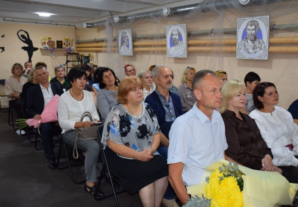Учасники заходу сидять у залі, прикрашеному портретами, трофеями та силуетом танцівниці, а на передньому плані лежить букет жовтих квітів.