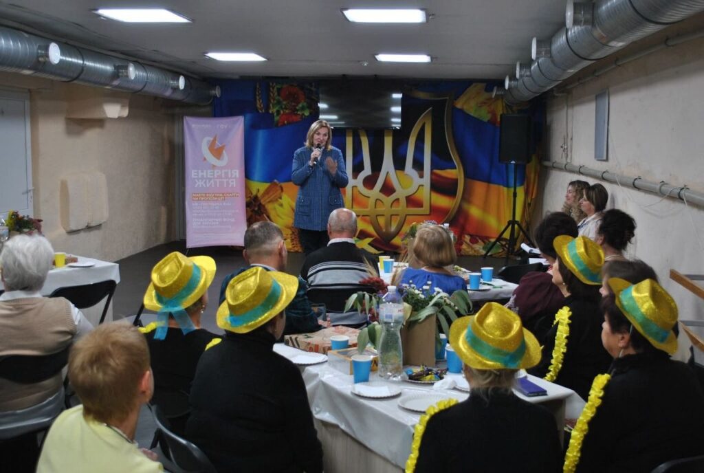 Жінка виступає з мікрофоном перед аудиторією в залі з прапором України та банером «Енергія життя».