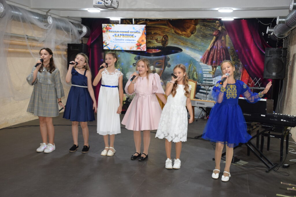 Шестеро дівчат у святкових сукнях співають у мікрофони на сцені під банером вокально-танцювального ансамблю «Барвінок».