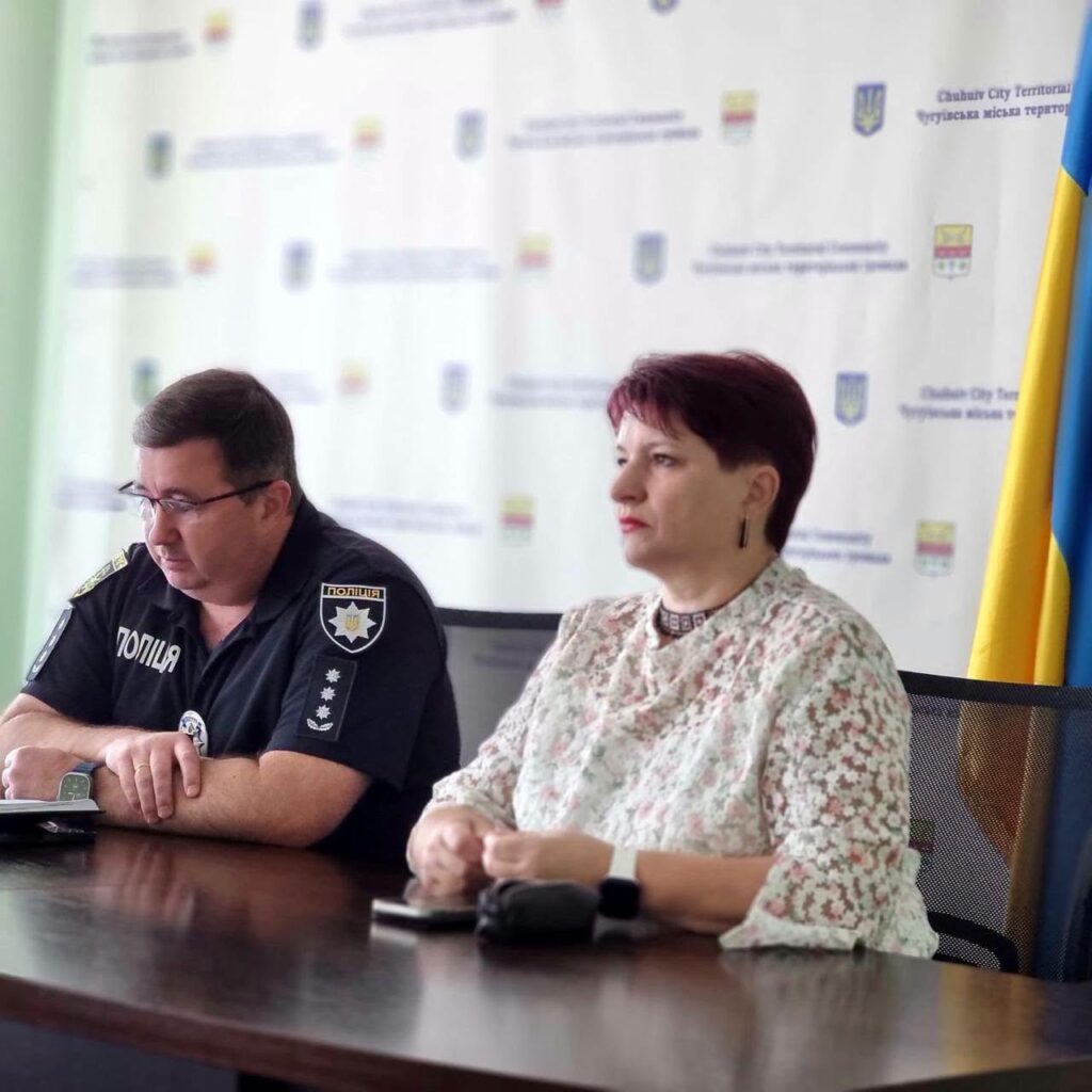 Поліцейський у формі та жінка в квітковій блузці сидять за столом під час офіційної зустрічі на тлі банера з написом «Чугуївська міська територіальна громада» та біля українського прапора.
