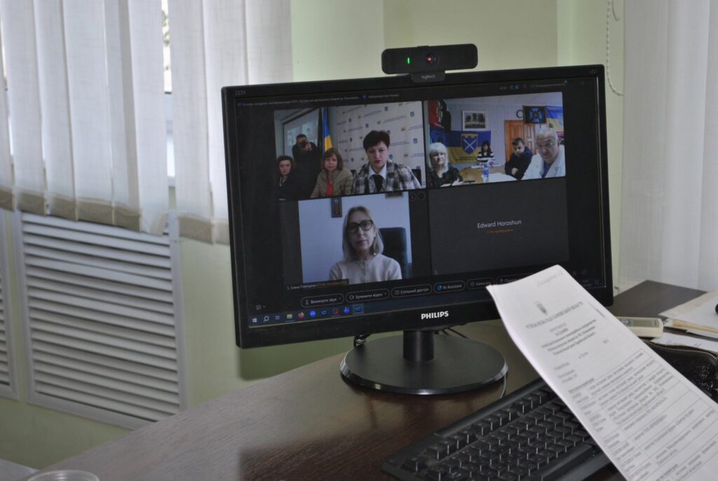 Комп'ютерний монітор Philips з підключеною веб-камерою відображає відеоконференцію з кількома учасниками, а на столі лежать документи та клавіатура.
