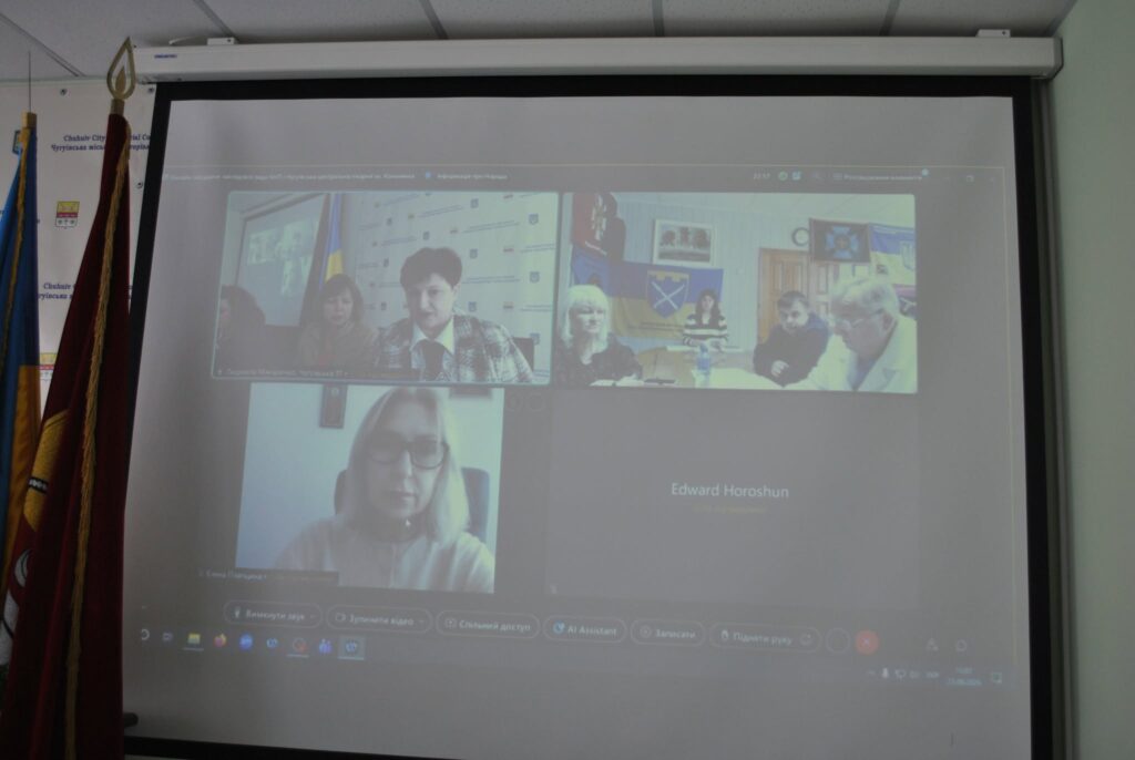 Проекторний екран відображає відеоконференцію з кількома учасниками, а поруч стоїть прапор.